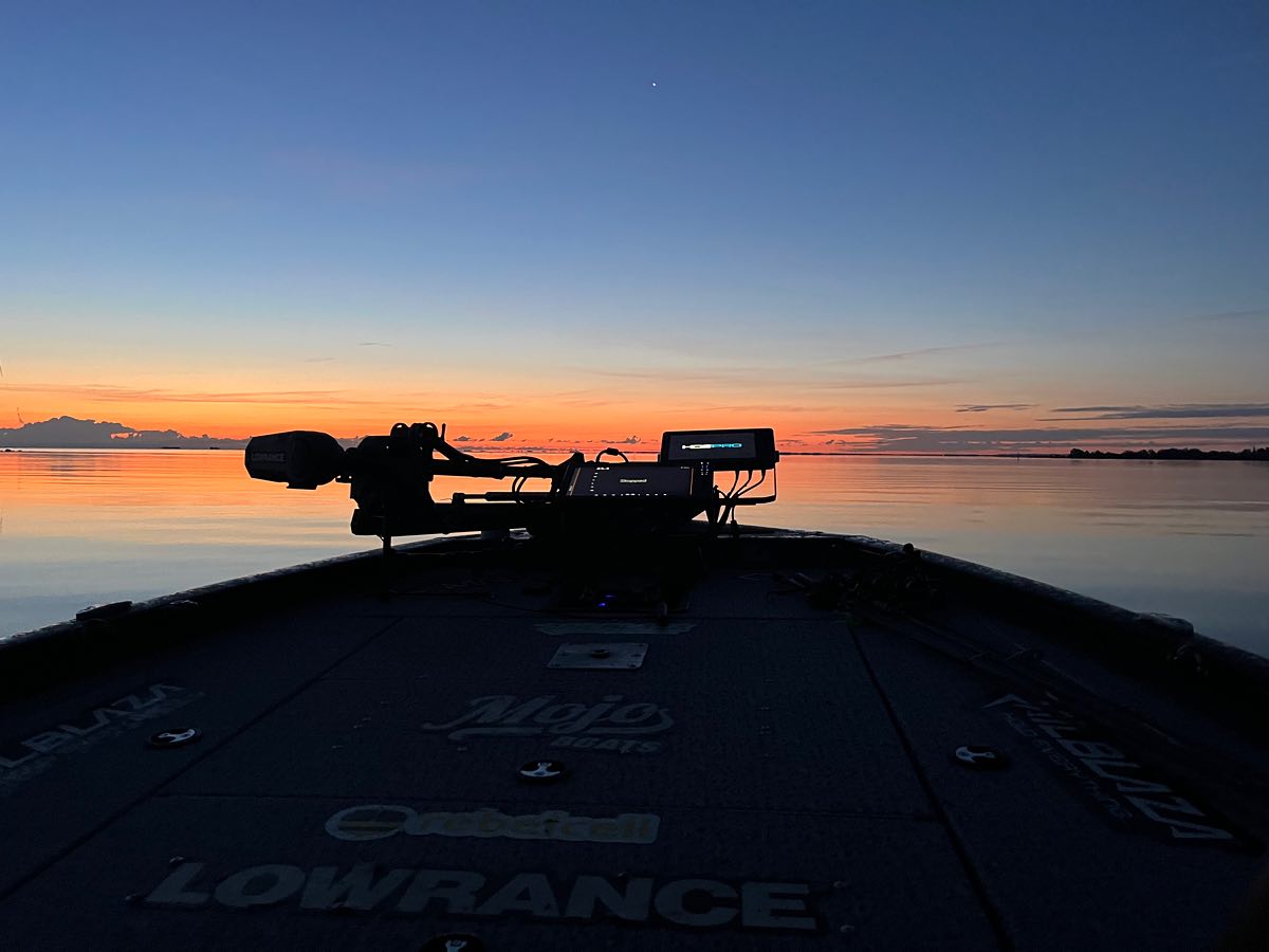 Solnedgång från fören av en båt på lugnt vatten, fiskeredskap i siluett och himlens färger speglas vid en rofylld fiskeresa.