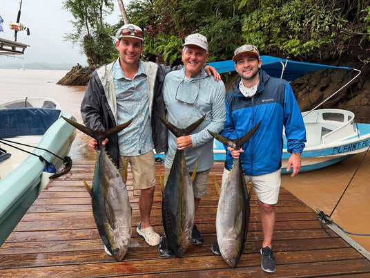 Tre sportfiskare på en brygga håller stora yellowfin-tonfiskar efter lyckad fiskeresa. Båtar och grönska i bakgrunden.