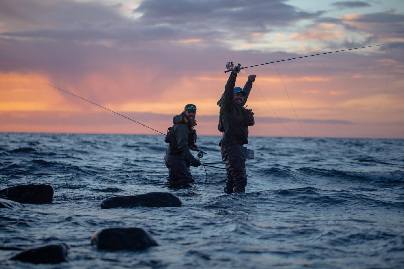 Två sportfiskare i fiskeutrustning står i havet vid solnedgången, med spö och leende, omgivna av vågor och klippor.