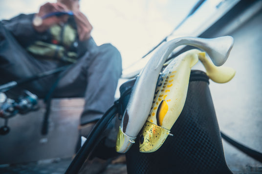 Närbild på två fiskformade beten fästa vid en fiskväska, suddig sportfiskare i båt med fiskeutrustning i bakgrunden.