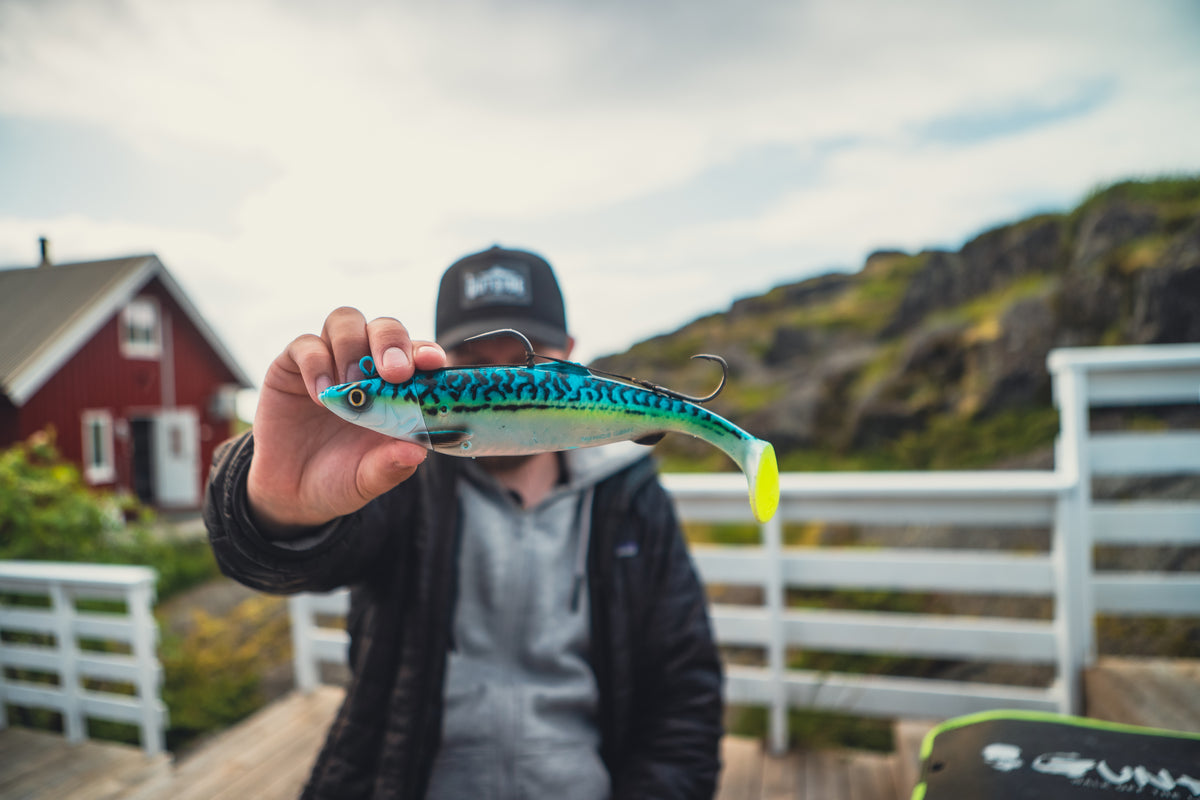 Sportfiskare i svart jacka och keps visar upp blå-gult fiskedrag. Röd stuga och klippor syns i bakgrunden.