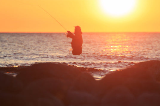 En siluett av en sportfiskare som kastar ut i havet vid solnedgång, solen speglar sig i vattnet. Inspiration för din fiskeresa.