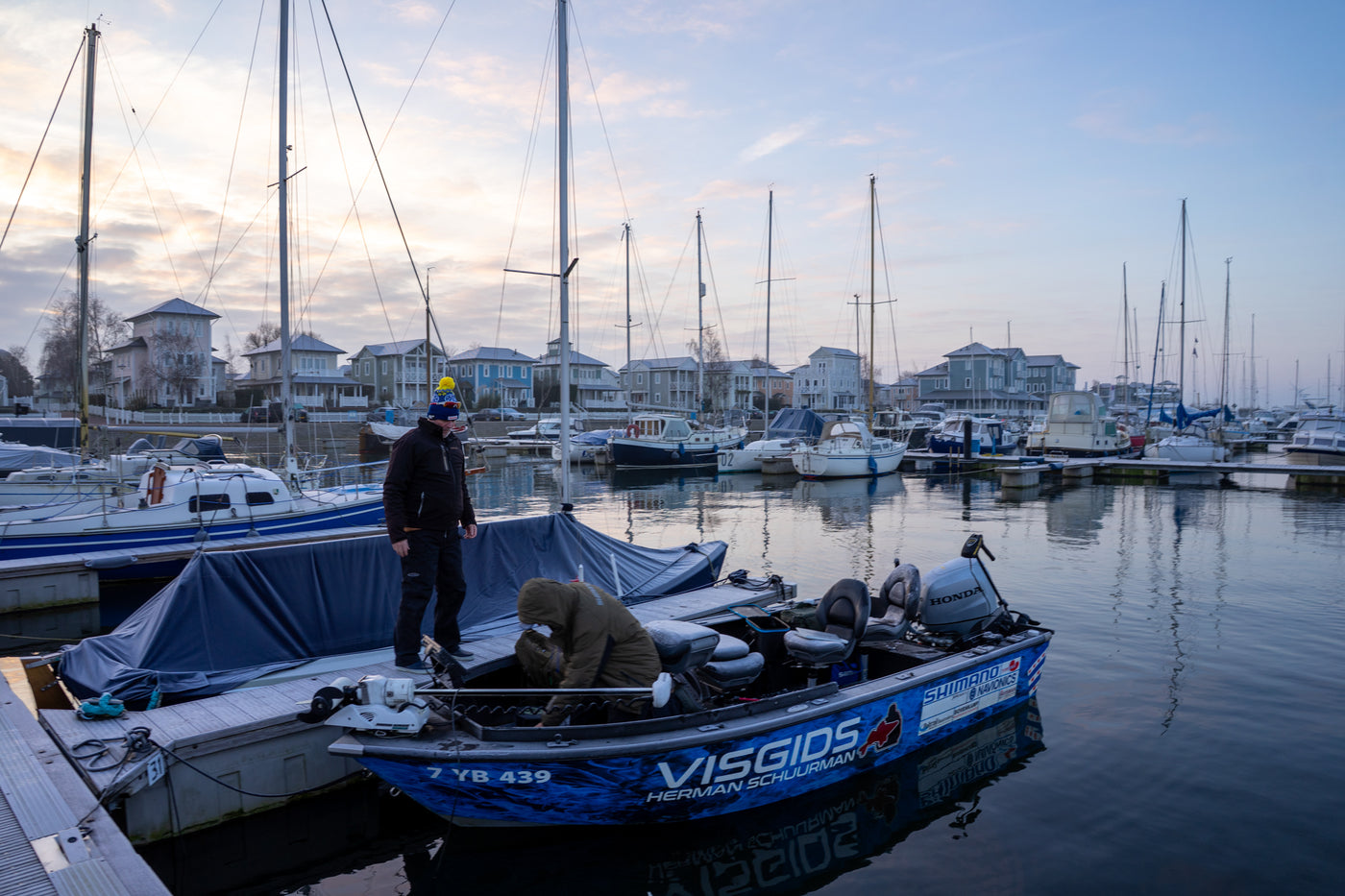 Två sportfiskare står på en liten blå fiskebåt vid bryggan, omgivna av segelbåtar och sjönära hus i gryning eller skymning.