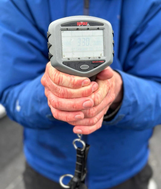En sportfiskare i blå jacka håller en digital våg som visar 3,30, med krok under - viktigt ögonblick på fiskeresan.