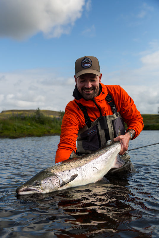 En man i orange jacka och keps står i grunt vatten och ler medan han håller en stor fisk med båda händerna under den blå himlen – en lyckad fisketur.