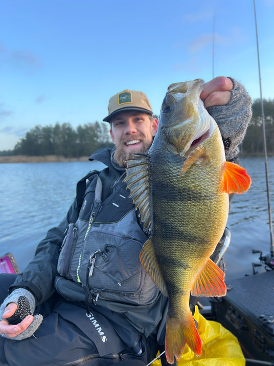 Leende man i fiskeutrustning sitter i båt på sjö, håller stolt upp stor gulgrön fisk. Sportfiske bland träd och blå himmel.