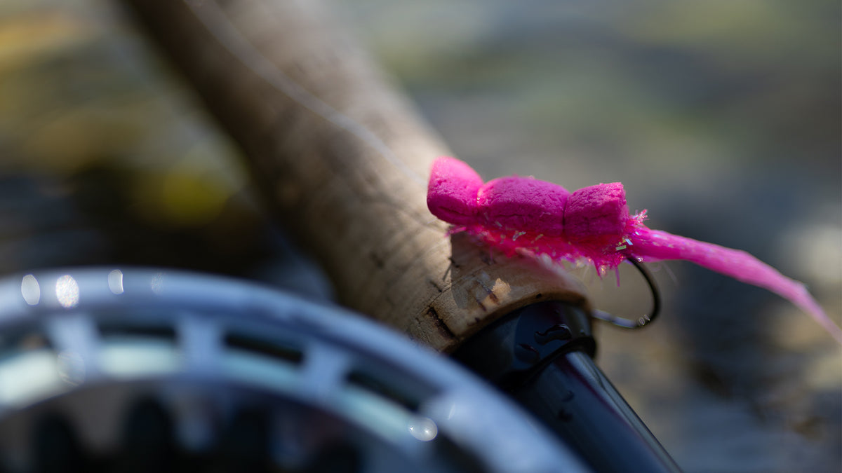 Närbild av fiskespö med rosa fluga vid vatten, perfekt för sportfiskare inför nästa fiskeresa; rullen syns suddigt i förgrunden.