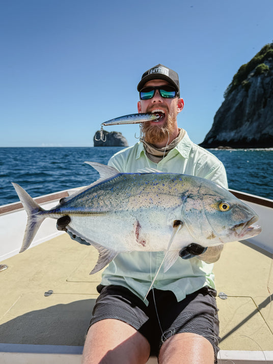 Sportfiskare på båt håller stor fisk med drag i munnen. Solglasögon, kepsar och handskar. Hav, klippor och blå himmel i bakgrunden.