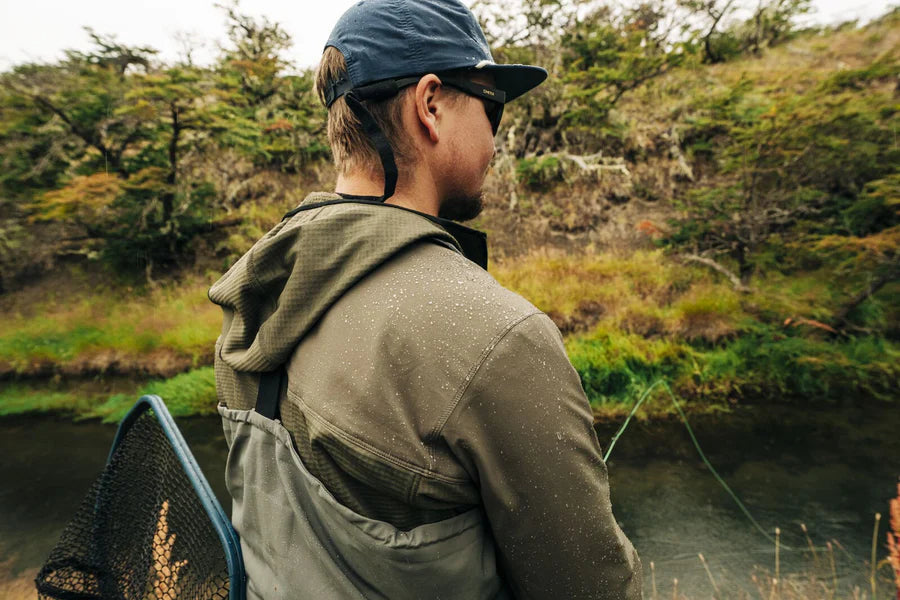 Person vid flod i Skwala Fishing Fusion Tactical Hoody, fisknät på ryggen, fiskeäventyr bland grönska och träd.