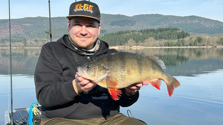 Sportfiskare i svart keps och hoodie håller en stor abborre i båten på sjön i Perchzilla