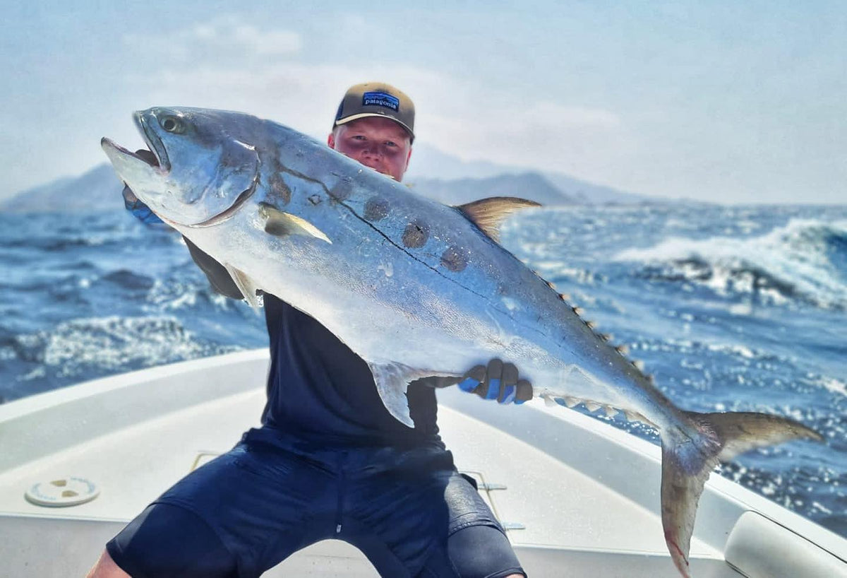 En sportfiskare i båt visar stolt upp en stor, silverglänsande fisk mot böljande hav och fjärran berg under klar himmel.