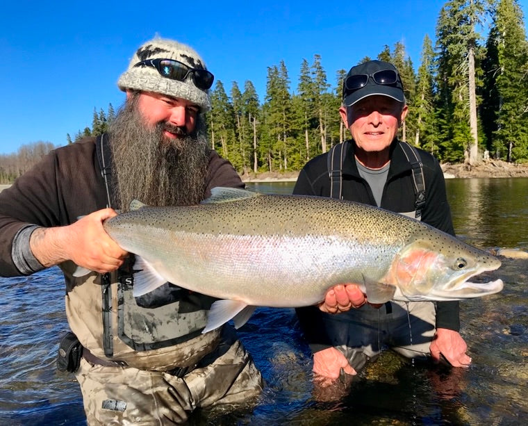 Två sportfiskare i fiskekläder står i grunt vatten, ler och håller en stor fisk. Tallskog och blå himmel syns bakom dem.