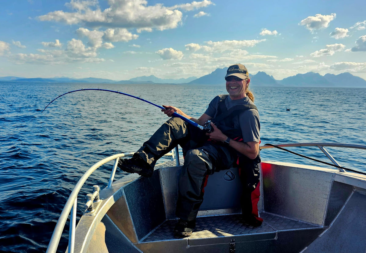 En sportfiskare sitter i båtens för, ler och håller ett fiskespö över blått vatten med fjäll och delvis molnig himmel i bakgrunden.
