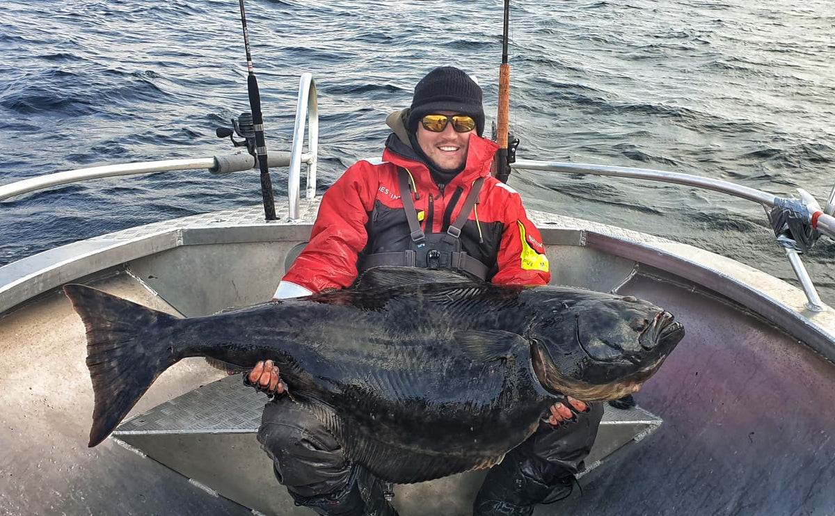 En sportfiskare i röd jacka och svart mössa sitter på båt, ler och håller en stor fisk med havet som bakgrund.