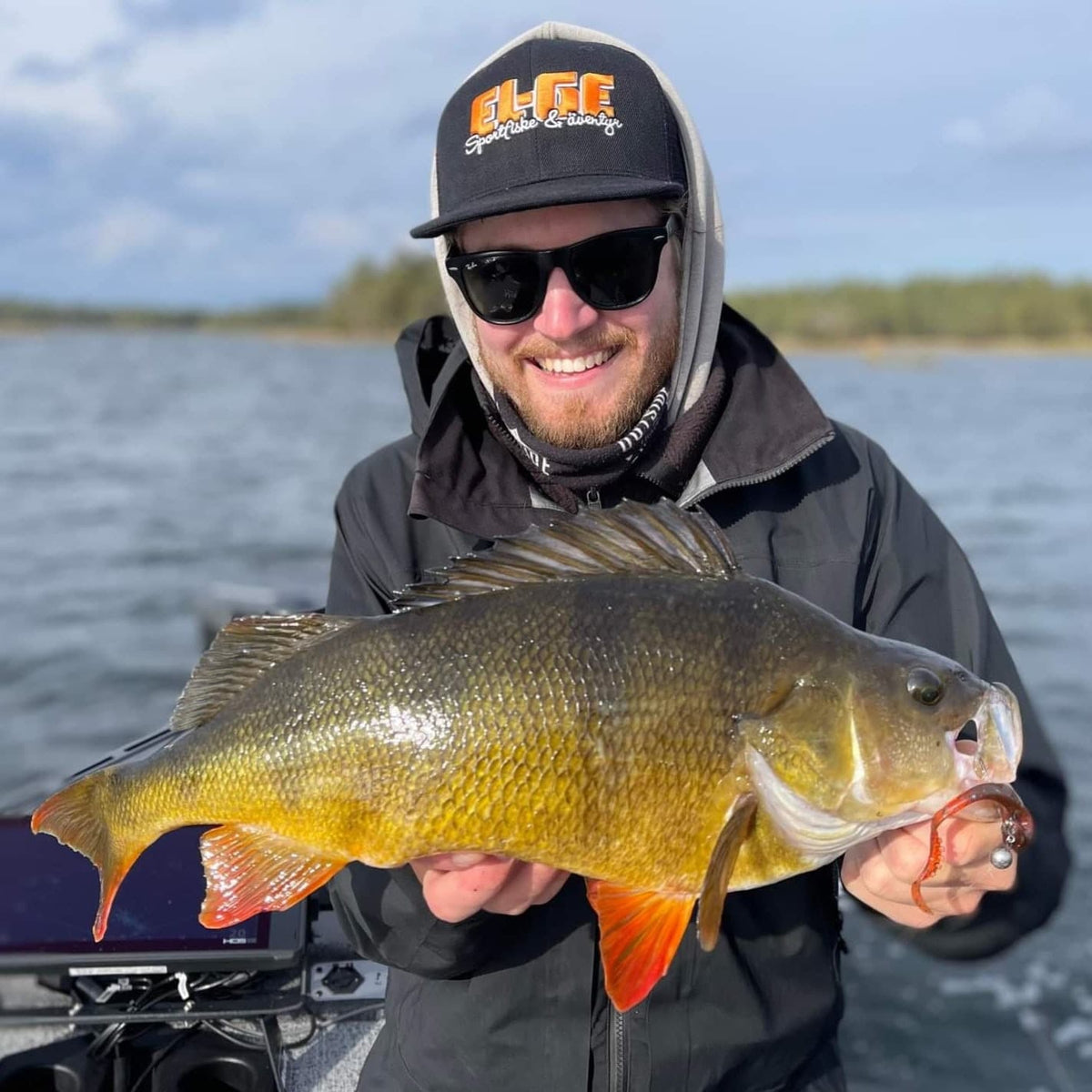 En sportfiskare med solglasögon, svart keps och jacka håller en stor färgglad fisk på båt, med vatten och kust i bakgrunden.