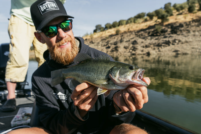 Skäggig sportfiskare med solglasögon och svart keps håller en largemouth bass i båt på lugn flod med klippstrand och träd.