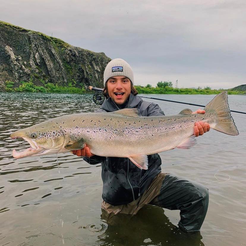Sportfiskare i vadare och mössa står leende i grunt vatten med stor fisk. Klippor och grönska vid äkta fiskeresa.