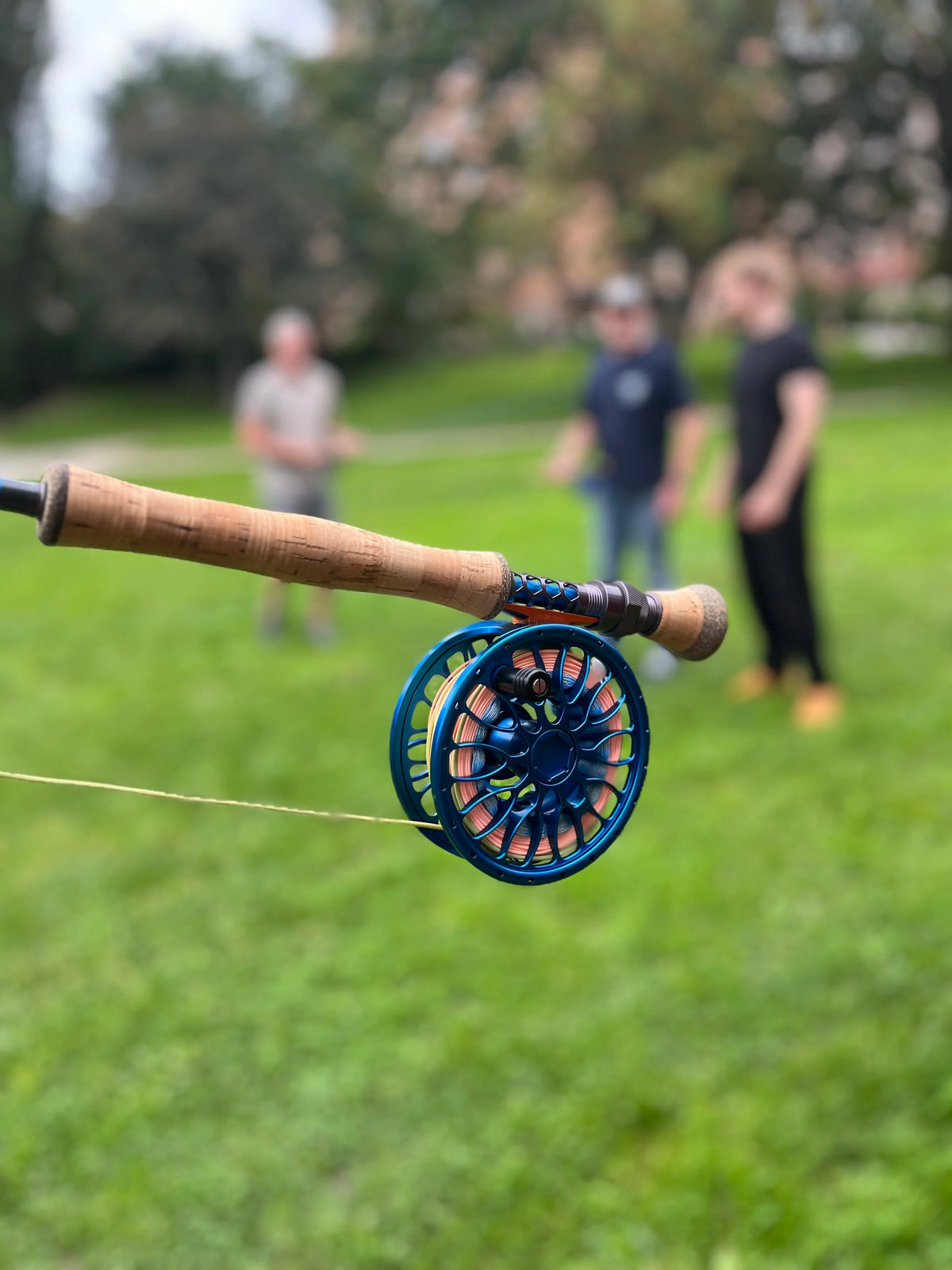 Närbild av en färgglad fiskerulle på ett spö, tre sportfiskare suddiga i bakgrunden på en grön äng.