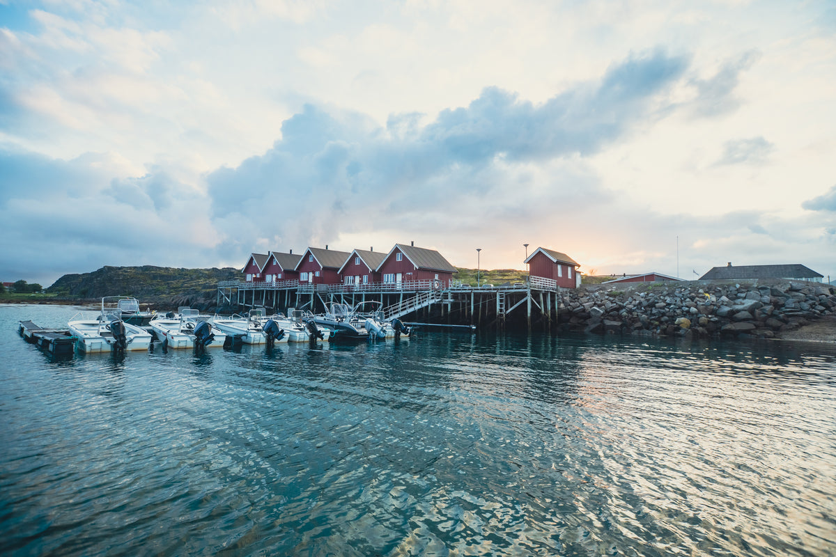 Röda trähus på pålar vid klippig kust, små fiskebåtar vid brygga, speglingar i vattnet - idealiskt för en fiskeresa.