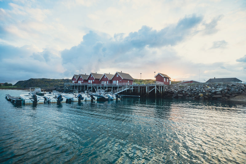 Röda trähus på pålar vid klippig kust, små fiskebåtar vid brygga, speglingar i vattnet - idealiskt för en fiskeresa.