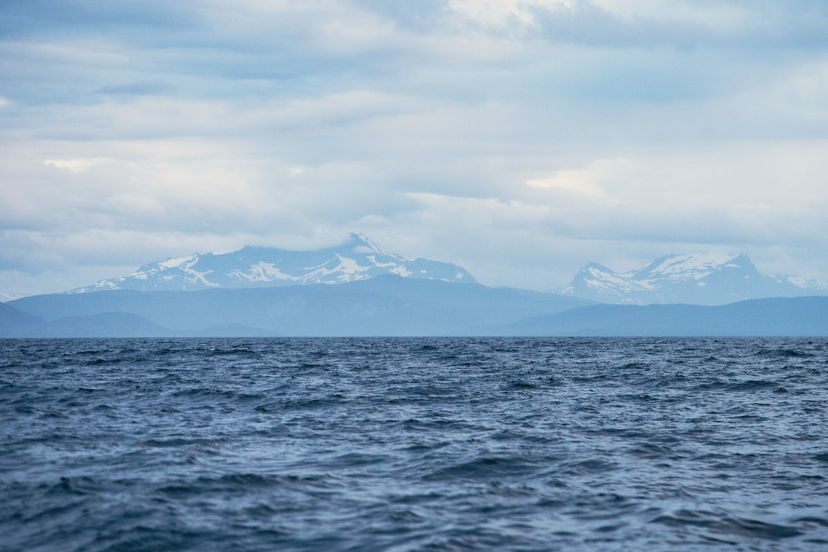 Stilla havsvatten i förgrunden, snötäckta berg och kullar i fjärran under molnig himmel - perfekt för sportfiskesan.
