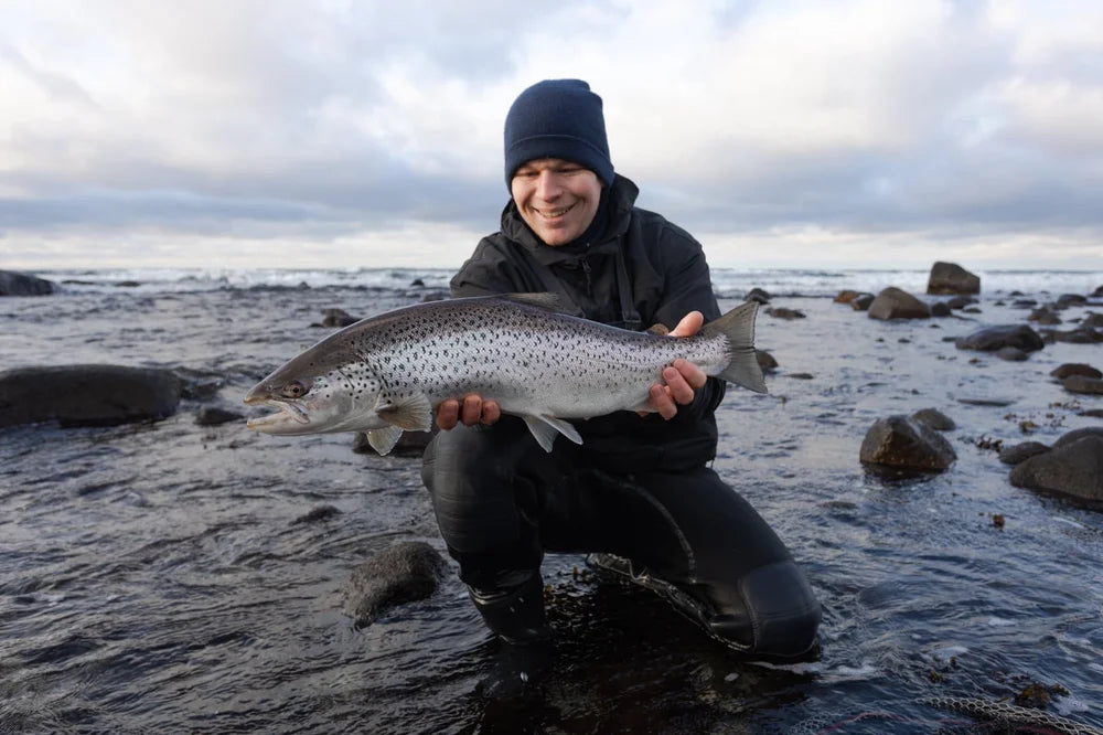 Sportfiskare i mörk jacka, mössa och vadare knäböjer vid stenig strand och håller en stor öring under molnig himmel på fiskeresa.