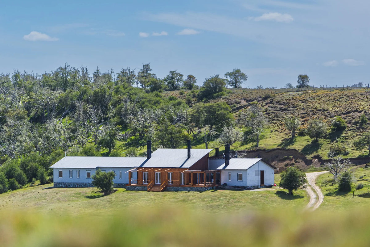 Rustik ranchvilla med tre sammanbyggda hus på gräsfält, perfekt för sportfiskare på fiskeresa. Kullar, skog och grusväg intill.
