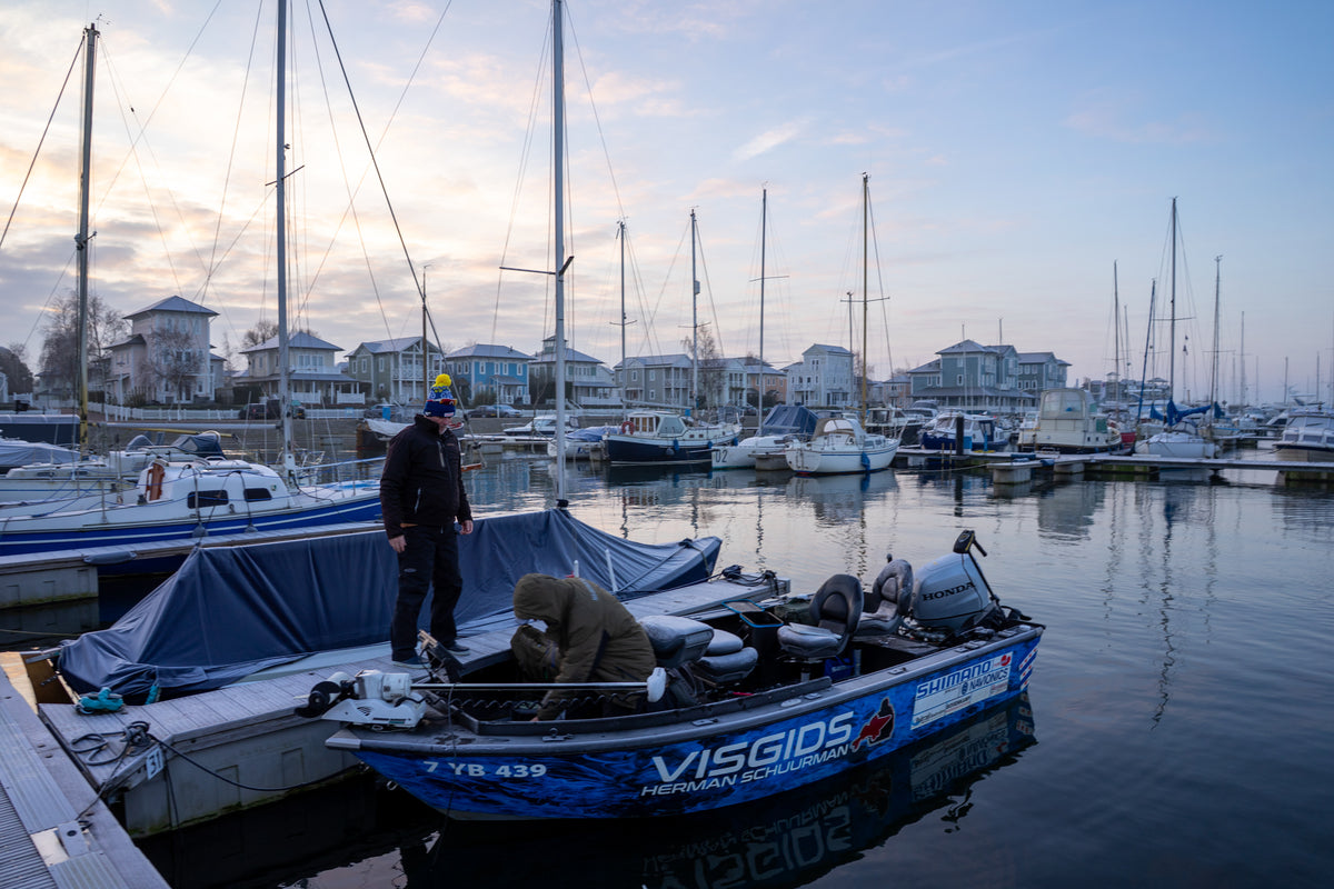 Två sportfiskare står på en liten blå fiskebåt vid bryggan, omgivna av segelbåtar och sjönära hus i gryning eller skymning.