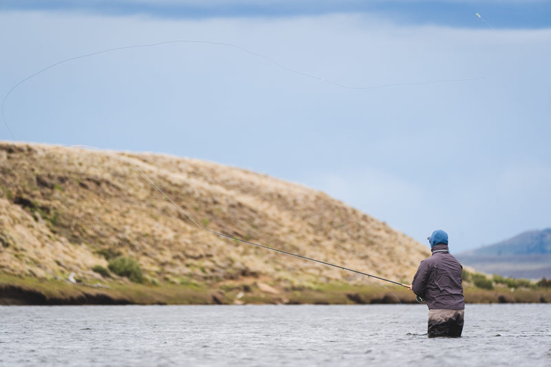 En person i blå keps och jacka vadar i en flod och flugfiskar under sin fiskeresa. Fiskelinan svävar genom luften med gröna kullar och en molnig himmel i bakgrunden – en dröm för alla fiskeentusiaster.