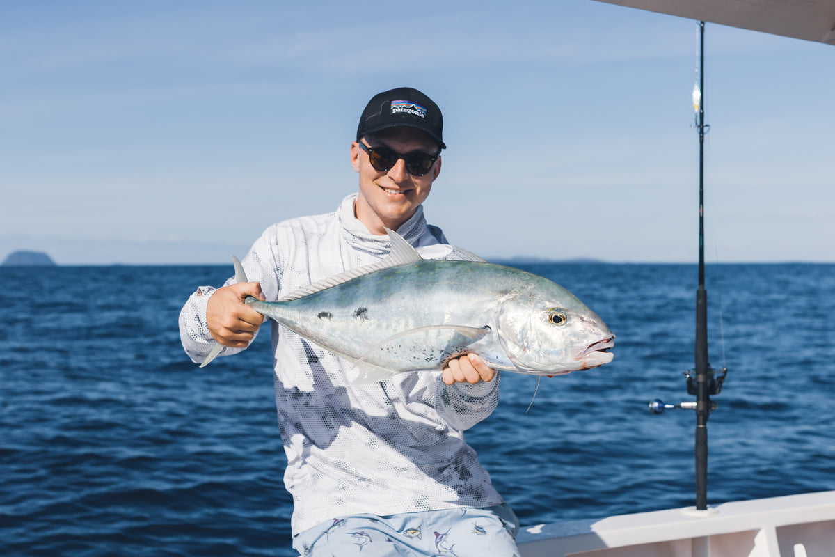 Sportfiskare med solglasögon och kepsar ler med stor fisk på båt under fiskeresa, hav och blå himmel i bakgrunden.