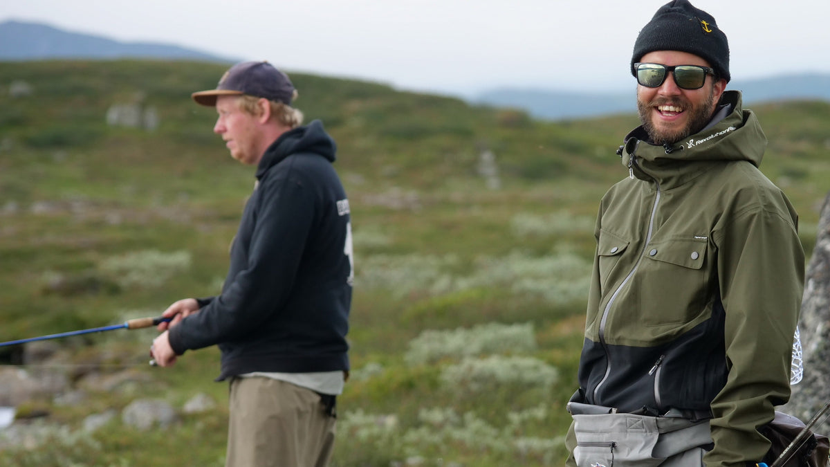 Två sportfiskare på fiskeresa: en fiskar i bakgrunden, en ler i förgrunden bland gräs under molnig himmel.
