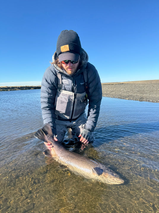 En person i friluftskläder och solglasögon sitter på knä i grunt vatten med en stor fisk – perfekt för en minnesvärd fiskeresa. Den klarblå himlen och det vidsträckta naturlandskapet skapar den bästa möjliga fiskeupplevelsen.