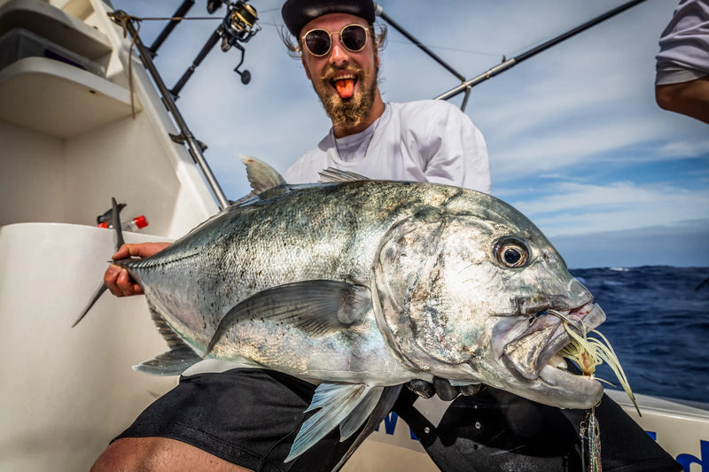 En person på fiskeresa håller en stor fisk ombord på båten, iklädd solglasögon och hatt. Havet och himlen syns i bakgrunden – ett fiskeäventyr när det är som bäst.