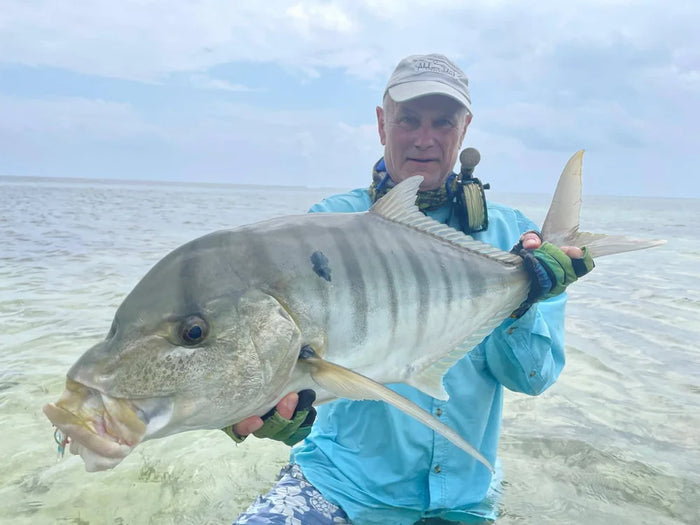 En sportfiskare i blå skjorta håller en stor silverglänsande fisk vid grunt vatten under molnig himmel på sin fiskeresa.