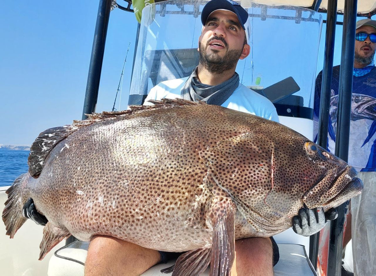 Man med keps och handskar visar stolt upp en stor prickig fisk i båt. Fiskekompis står bakom med spön under blå himmel.
