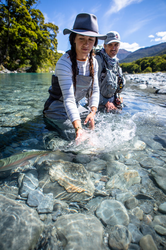 Kvinna i vadarbyxor släpper tillbaka fisk i klar älv på fiskeresa, medan man i friluftskläder ser på. Natur och blå himmel.